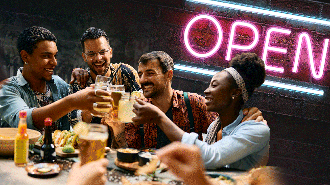 A group of friends eating and drinking at a restaurant, with a neon pink 'open' sign flashing in the background