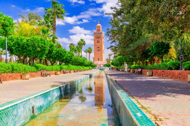 The minaret of the Koutoubia Mosque in Marrakesh, Morocco, at the end of a waterway in the pavement and framed by bright green trees.