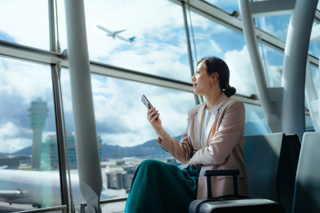 Confident young businesswoman with suitcase looking ahead while using smartphone waiting for her flight at airport terminal.