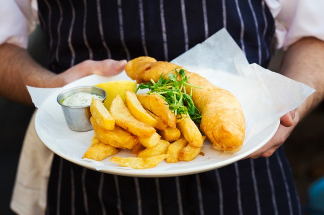 A chef holds a dish of fish and chips with garnish and tartare sauce.