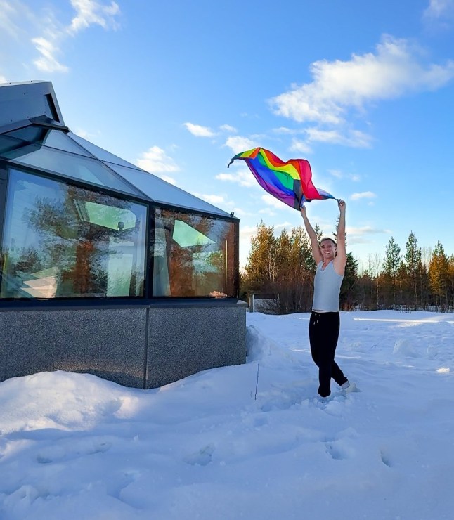 Calum holds a Pride flag in the breeze outside in the snow in Rovaniemi, Finland. There is a hotel room to the left of the photo and the sky is blue.