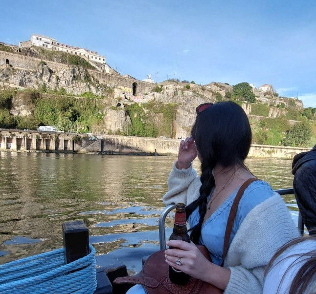 A woman looks out over the Douro River in Porto, up at a castle on a sunny day. She has her back to the camera and is holding a drink.