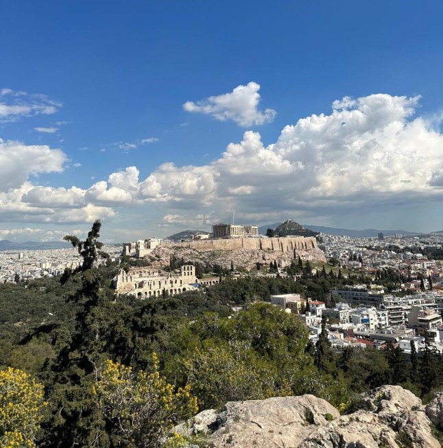 A distant view of the Acropolis on a hilltop in Athens, Greece on a sunny day. The sky is blue and cloudy.