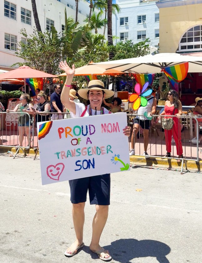 A woman holds a sign that reads 'Proud mother of a transgender son' on a sunny street in Miami at Pride