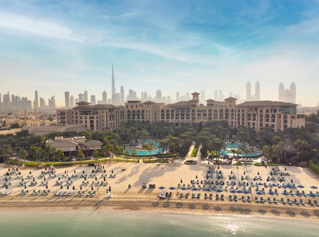 An aerial view of the beachfront and swimming pools at the Four Seasons resort in Dubai, with the city skyline in the background on a sunny day.