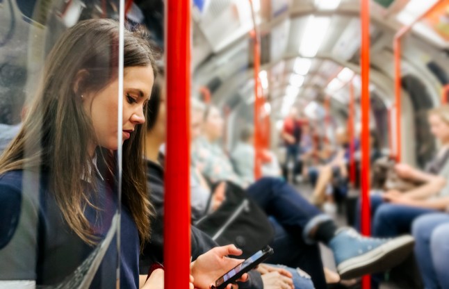 A young woman on a London Underground train looking at her phone, with someone crossing their legs a few seats away.