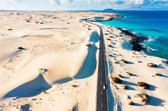 A stunning road in Fuerteventura, surrounded by sand dunes on one side and turquoise blue sea on the other.