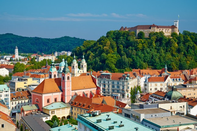A cityscape view of Ljubljana, Slovenia, showing colourful buildings, the Franciscan church, Saint Nicholas church and the Castle on a tree-covered hill.