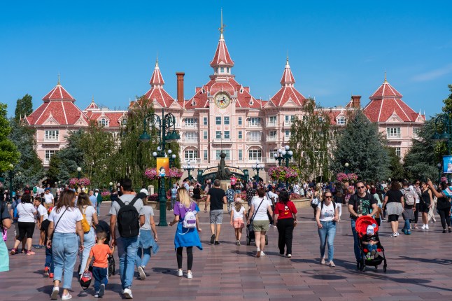 People walk in front of the entrance to Disneyland Paris in France on a sunny day, with blue sky