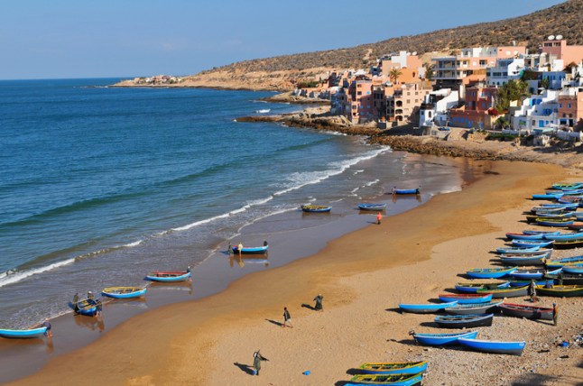 A sunny view of a beach in Taghazout, Morocco, where the golden sand is lined with blue boats.
