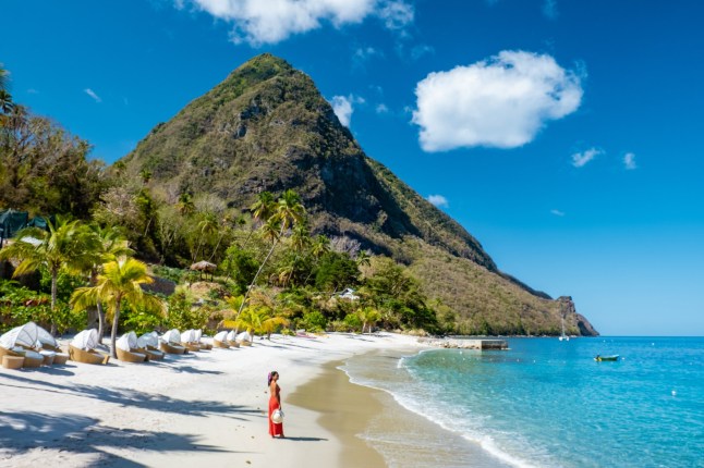 A woman standing on the white sandy beach in St Lucia, with a large rock formation, palm trees and blue ocean surrounding her.