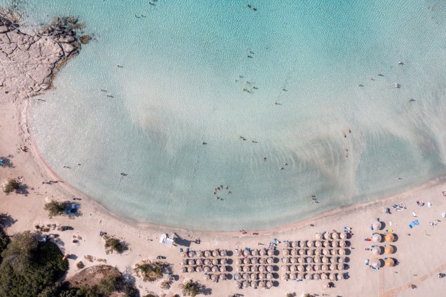 Sandy beach with umbrella in Elafonisi, Crete, Greece. Aerial drone view of wavy turquoise sea water, pink sand background. Famous summer resort.